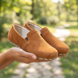 May include: A pair of tan suede slip-on shoes featuring a rounded toe, a low-cut collar, and a visible stitched detail along the edge of the sole. The interior lining is light-colored, and the exterior is a solid, matte tan suede material.