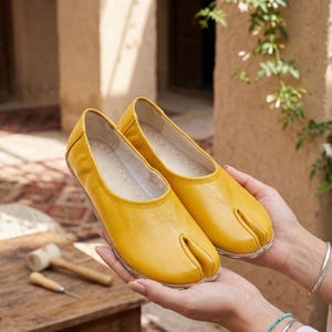 Op de afbeelding: Een paar felgele leren schoenen met split-teen. De schoenen hebben een ronde neus en een apart compartiment voor de grote teen. De zolen zijn van een doorschijnend materiaal. De schoenen worden op een houten oppervlak gepresenteerd.