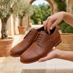 May include: A pair of brown leather dress shoes with a cap toe and lace-up closure. The shoes are held in a person's hands, showcasing their polished appearance. The shoes have a classic design, suitable for formal or business attire.
