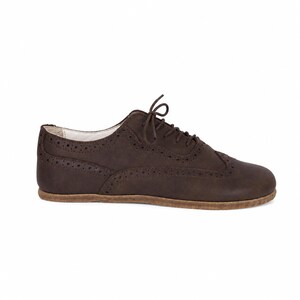 May include: A dark brown lace-up oxford shoe featuring decorative brogue perforations along the side panels and toe cap. The shoe has a low-profile design with a flat, textured tan sole and a light-colored interior lining visible at the collar.