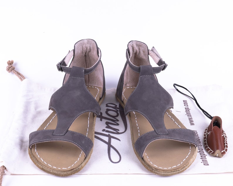Puede incluir: Un par de sandalias de cuero gris con correas de tobillo y suela de cuero marr&oacute;n. Las sandalias est&aacute;n sobre una superficie blanca con una bolsa de cord&oacute;n blanco y una etiqueta de cuero marr&oacute;n.