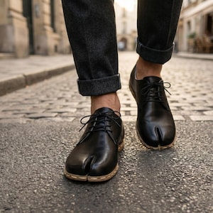 Op de afbeelding: Zwarte leren schoenen met split-teen en een vetersluiting. De schoenen hebben een uniek split-teen ontwerp en een lichtgekleurde zool. De persoon draagt een donkergrijze broek met opgerolde manchetten, lopend op een geplaveide straat.