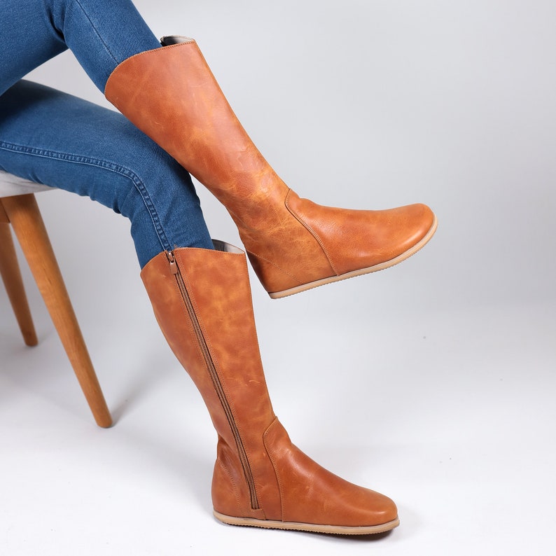 May include: Tan leather knee-high boots with a side zip. The boots have a flat sole and are worn with blue jeans. The leather has a slightly distressed look. The boots are approximately 40 cm in height.