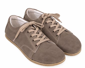 May include: A pair of low-top sneakers in a muted taupe color featuring a smooth matte finish, matching taupe laces, metal eyelets, and a light tan textured sole. The interior lining is white, and the design includes visible tonal stitching.