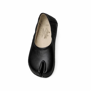 May include: Black leather split-toe ballet flats. The shoes have a rounded toe and a split design, with a small heel. The sole is black, and the interior is a light beige color. The shoes are designed for comfort and flexibility.