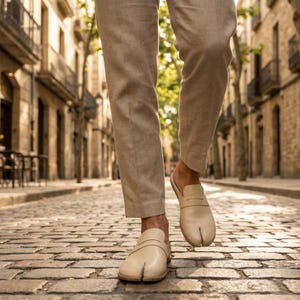 May include: Cream-colored leather split-toe shoes with a strap across the top. The shoes are worn with beige linen pants. The image is taken on a cobblestone street with buildings in the background.