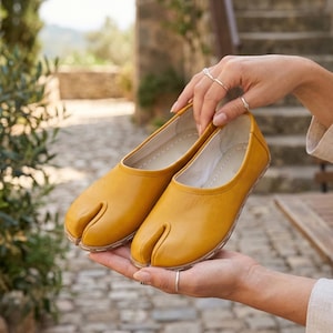 May include: A pair of mustard yellow split-toe flats. The shoes are made of smooth leather and have a unique design with a separate space for the big toe. The flats are held in a person's hands, showcasing their color and style.