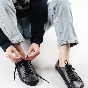 May include: A person is tying the laces of a pair of black leather low-top sneakers. The sneakers have a white rubber sole and black laces. The person is wearing blue jeans and a dark blue sweatshirt.