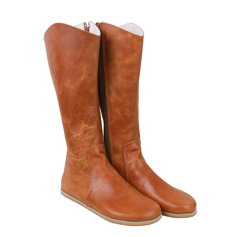 May include: A pair of tall, knee-high brown leather boots. The boots have a side zip and a flat sole. The leather has a distressed appearance with visible texture and colour variations. The interior lining is white.