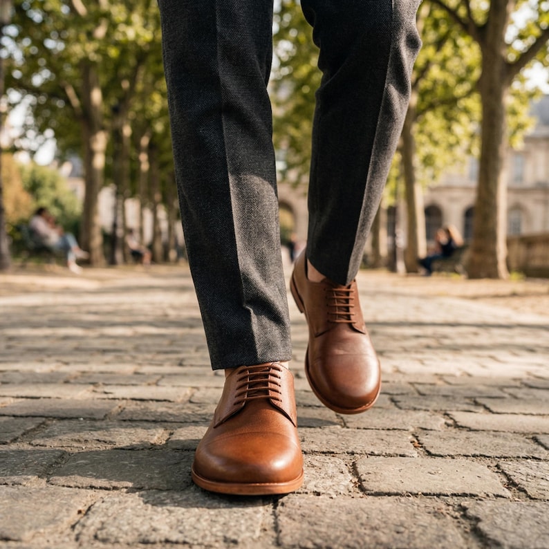 May include: A person walking in brown leather dress shoes with a lace-up design. The shoes are a warm brown color and are paired with gray trousers. The image is taken outdoors on a cobblestone path.