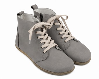 Op de afbeelding: Een paar enkelhoge laarzen in een matte grijze afwerking met lichtbeige veters, metalen oogjes, een ritssluiting aan de zijkant en een treklipje bij de hiel. De laarzen hebben een ronde neus en een platte zool met een textuurrand.
