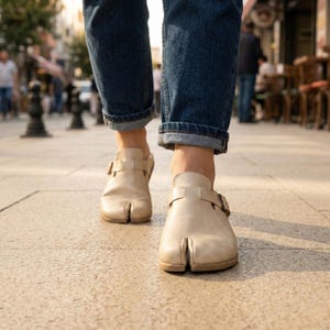 May include: Beige leather split-toe clogs with buckle straps. The clogs are worn with blue denim jeans, cuffed at the ankles. The image is taken from a low angle, showing the clogs on a stone walkway.