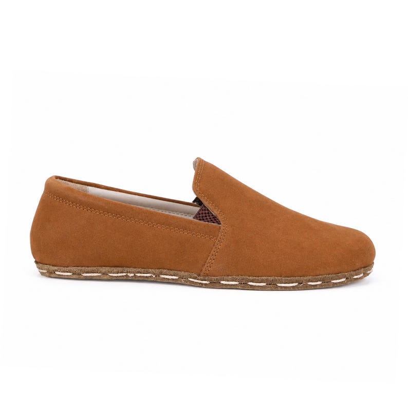 May include: A tan suede slip-on shoe featuring a rounded toe, a low-cut collar, and a visible thick brown sole with prominent light-colored stitching along the entire perimeter of the base, showcasing a clean and simple profile design.