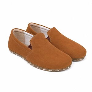 May include: A pair of tan slip-on shoes featuring a smooth suede-like exterior, a rounded toe, and a light-colored interior lining. The shoes have a visible stitched detail along the base where the upper meets the sole, with a small brown patterned fabric accent at the side opening.