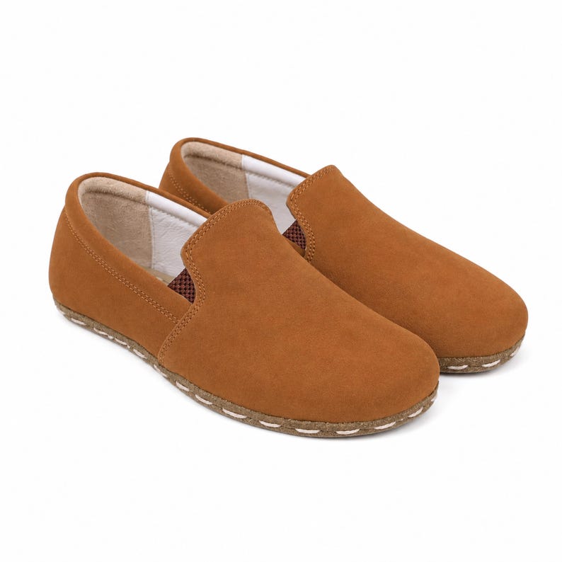 May include: A pair of tan slip-on shoes featuring a smooth suede-like exterior, a rounded toe, and a light-colored interior lining. The shoes have a visible stitched detail along the base where the upper meets the sole, with a small brown patterned fabric accent at the side opening.