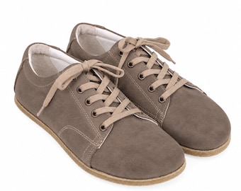 May include: A pair of low-top sneakers in a muted taupe color with a smooth, matte finish. These shoes feature matching taupe laces, metal eyelets, white interior lining, and a light tan textured rubber sole with visible stitching along the edge.