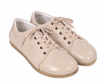 May include: A pair of low-top sneakers in a light beige color. These shoes feature a smooth leather-like upper, matching beige laces threaded through metal eyelets, a rounded toe box, and a flat, textured brown rubber sole for traction.