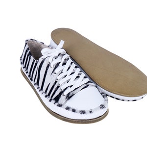 May include: A pair of white canvas sneakers with black zebra print on the upper. The laces are white and the shoes have a brown rubber sole.