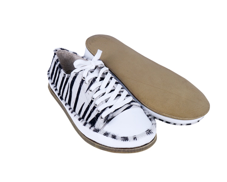 May include: A pair of white canvas sneakers with black zebra print on the upper. The laces are white and the shoes have a brown rubber sole.