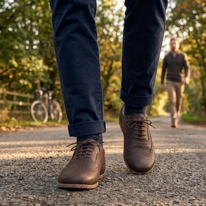 May include: A pair of dark brown leather oxford shoes with brogue detailing, featuring a rounded toe, a lace-up closure, and a low heel. The shoes are worn with dark blue cuffed trousers and dark socks, showcasing a classic footwear design.