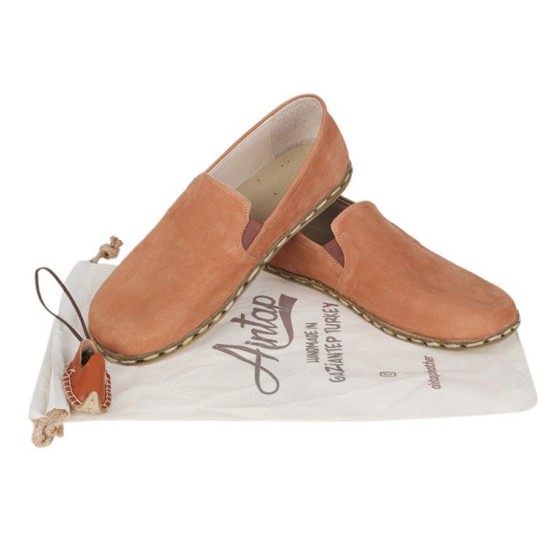 May include: A pair of tan leather slip-on shoes with visible stitching along the soles, resting on a white fabric drawstring bag printed with the text Aintap, Handmade in Gaziantep Turkey, and a miniature leather shoe keychain attached.