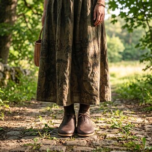 May include: A long, pleated, olive-green skirt with a dark, intricate botanical print, paired with brown socks and dark brown leather oxford shoes featuring brogue detailing. A small, woven, light-brown wicker basket is held at the side.