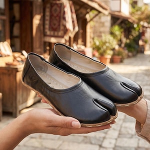 Op de afbeelding: Een paar zwarte leren schoenen met split-teen. De schoenen hebben een ronde neus en een apart compartiment voor de grote teen. De zolen zijn lichtbruin van kleur en de schoenen worden in de handen van een persoon gehouden. De achtergrond is een vage buitenscène.