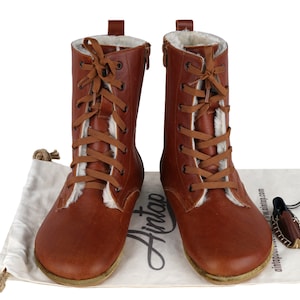 May include: Brown leather boots with a lace-up front and a zip on the side. The boots have a white fur lining and are sitting on a white fabric surface with a brown leather bag and a brown leather tag that says "Hintap" on it.