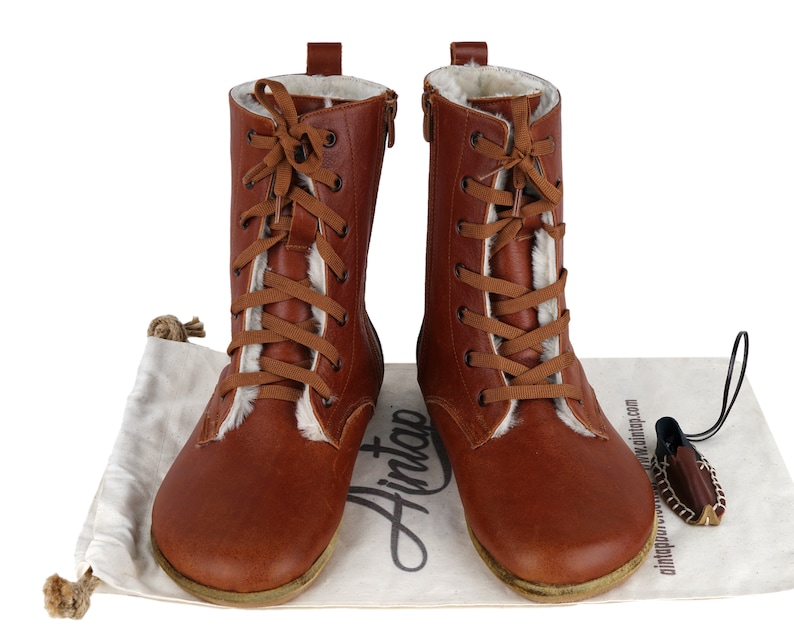 May include: Brown leather boots with a lace-up front and a zip on the side. The boots have a white fur lining and are sitting on a white fabric surface with a brown leather bag and a brown leather tag that says "Hintap" on it.