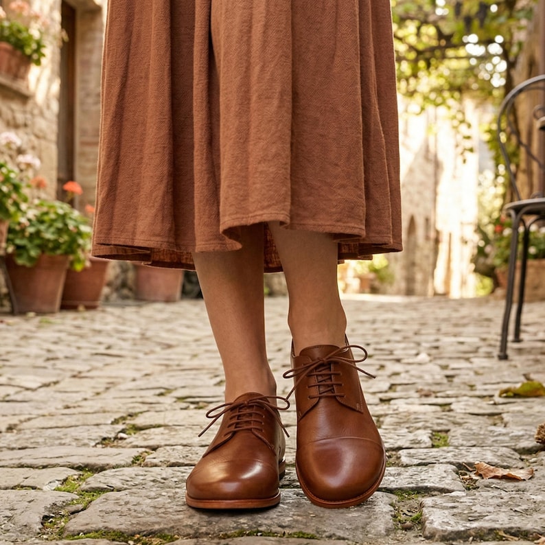 May include: Brown leather lace-up shoes. The shoes have a classic design with a rounded toe and a low heel. The laces are tied in a neat bow. The shoes are worn with a brown dress. The background is a cobblestone street.