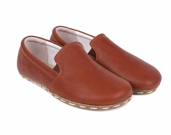 May include: A pair of slip-on shoes in a smooth, medium-brown leather finish. The shoes feature a rounded toe, a low-cut collar, and a light-colored interior lining. A thick, textured tan sole is attached to the upper with visible white stitching.