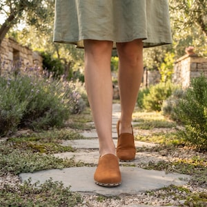 May include: A pair of tan suede slip-on shoes with a rounded toe and visible white stitching along the sole edge. The shoes feature a low-cut topline and a flat, flexible construction, worn with a light olive green knee-length skirt.