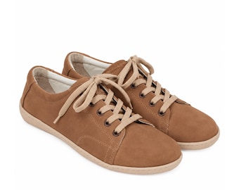 May include: A pair of low-top sneakers in a solid tan color. These shoes feature a smooth matte finish, matching tan laces threaded through dark metal eyelets, and a light beige rubber sole. The interior lining is a contrasting off-white color.