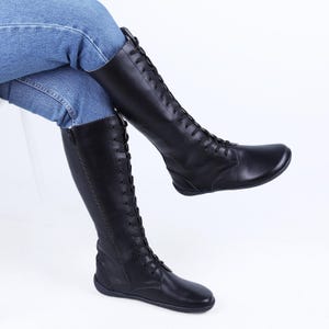 Könnte beinhalten: Schwarze kniehohe Stiefel mit Schnürung und seitlichem Reißverschluss. Die Stiefel sind aus glattem Material und haben eine flache Sohle. Die Stiefel werden mit einer blauen Jeans getragen.