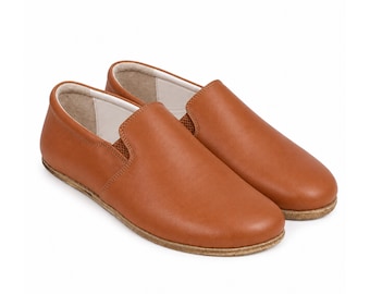 May include: A pair of slip-on shoes in a smooth tan leather finish. These shoes feature a rounded toe, a low-cut collar with visible elastic side gussets for flexibility, and a flat, textured brown sole. The interior lining is a light cream color.