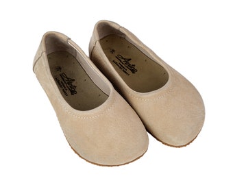 Puede incluir: Un par de bailarinas de ante beige con punta redonda y suela plana. Los zapatos tienen un forro de cuero y una plantilla de cuero. El nombre de la marca "Antaea" está impreso en la plantilla.