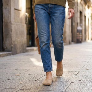 Op de afbeelding: Een persoon loopt op een geplaveide straat met een blauwe spijkerbroek, een bruine trui en beige schoenen met split-teen. Een bruine leren tas wordt in de linkerhand gedragen. De schoenen hebben een uniek ontwerp met een scheiding voor de grote teen.