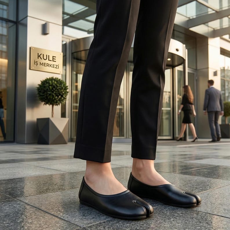 May include: Black leather split-toe ballet flats. The shoes have a rounded toe and a low heel. The image shows the shoes on a person's feet, with a business building in the background.