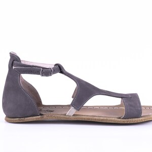 Puede incluir: Sandalias de cuero gris con una tira en T y cierre de hebilla. Las sandalias tienen una suela plana y un borde cosido.