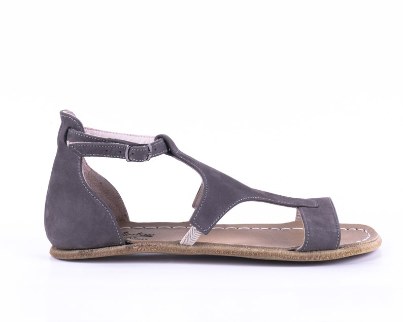 Puede incluir: Sandalias de cuero gris con una tira en T y cierre de hebilla. Las sandalias tienen una suela plana y un borde cosido.