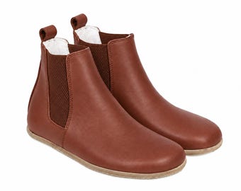 Könnte beinhalten: Ein Paar braune Stiefeletten mit glatter Oberfläche, abgerundeten Zehenpartien und dunkelbraunen elastischen Seiteneinsätzen. Jeder Stiefel verfügt über eine kleine Zugschlaufe an der Ferse und eine helle, flache Sohle. Das Innenfutter ist weiß.