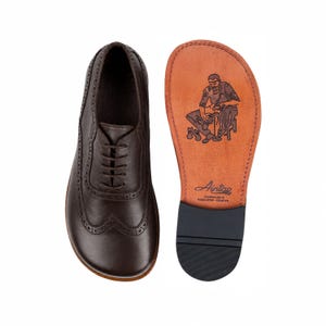 Può includere: Una scarpa oxford in pelle marrone scuro con dettagli brogue e lacci, abbinata alla sua suola. La suola presenta un'illustrazione stampata di un calzolaio, il marchio Aintap e il testo Handmade in Gaziantep-Türkiye.
