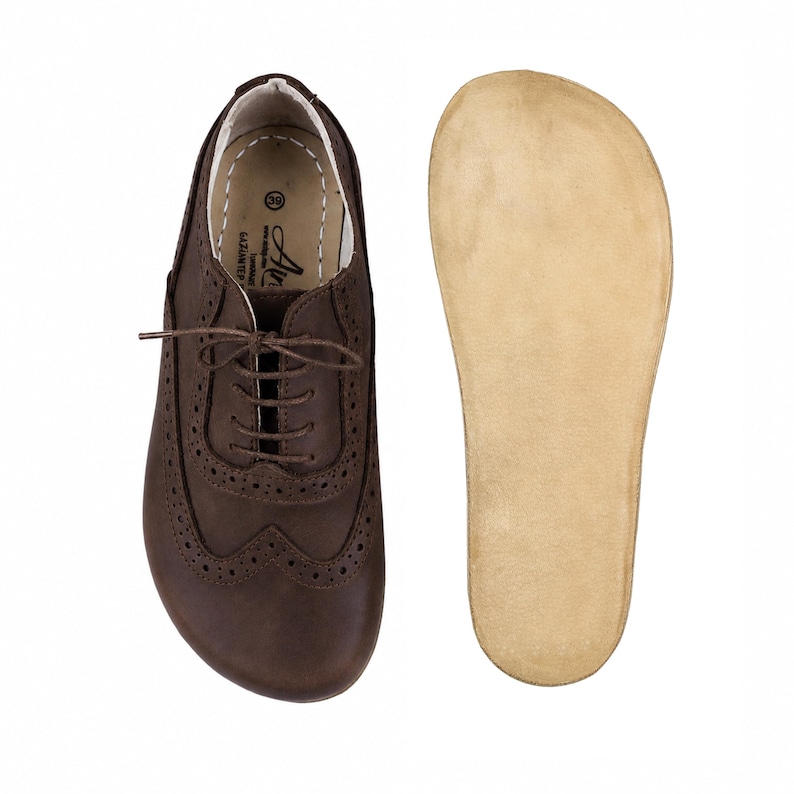 May include: A dark brown leather oxford shoe with brogue detailing, thin brown laces, and a size 39 label inside, paired with a separate, smooth, light beige leather insole. The shoe features a rounded toe and visible stitching along the collar.