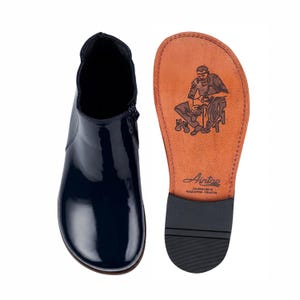 May include: A dark navy blue patent leather ankle boot with a side zipper and a tan leather sole. The sole features a black rubber heel cap and a stamped illustration of a shoemaker at work with the text Aintap Handmade in Gaziantep-Türkiye.
