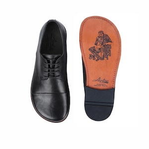 May include: Black leather dress shoe with a lace-up design, next to a shoe sole with a brown leather insole. The insole features a craftsman illustration and the brand name "Aintoe". The shoe is designed for comfort and style.