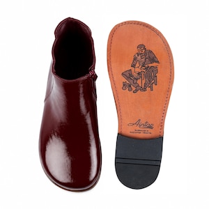 May include: A glossy dark burgundy ankle boot with a side zipper and a tan leather sole. The sole features a stamped illustration of a shoemaker at work, the brand name Aintap, and the text Handmade in Gaziantep-Türkiye with a black heel.