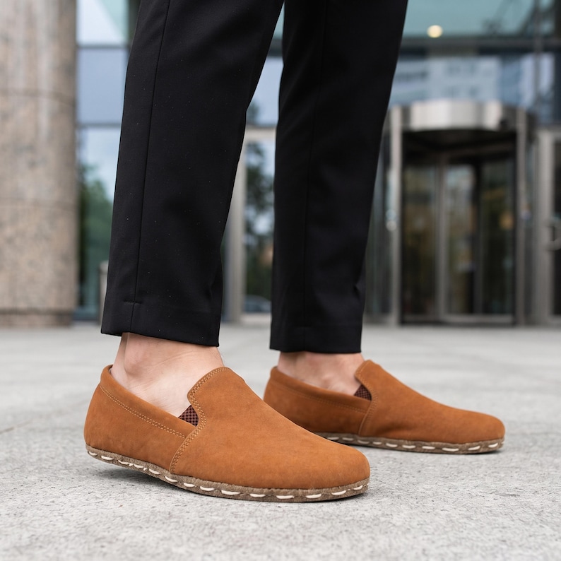 May include: A pair of tan suede slip-on shoes featuring a rounded toe, a low-profile silhouette, and a visible light-colored stitched detail along the edge of the sole. The interior lining shows a small brown and white checkered pattern.