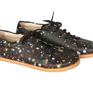 Op de afbeelding: Een paar zwarte leren sneakers met een kleurrijk verfspatten ontwerp. De sneakers hebben witte veters en een bruine rubberen zool.