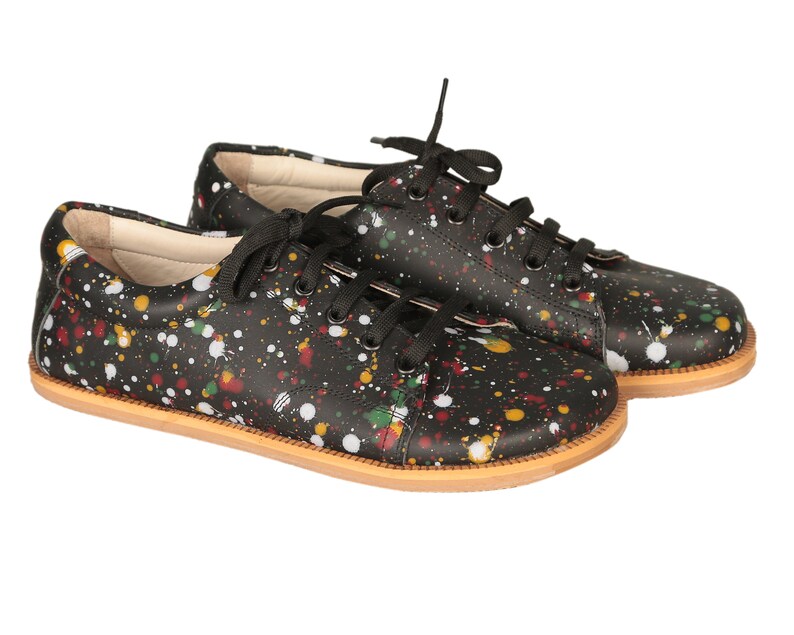 Op de afbeelding: Een paar zwarte leren sneakers met een kleurrijk verfspatten ontwerp. De sneakers hebben witte veters en een bruine rubberen zool.