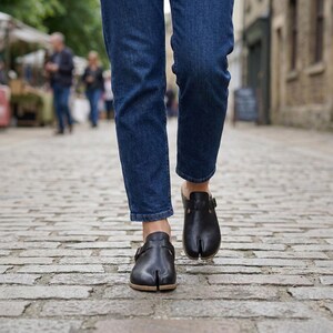 May include: Black leather split-toe clogs with a buckle closure. The clogs have a wooden sole and are being worn with blue jeans. The shoes are designed with a split between the big toe and the other toes.